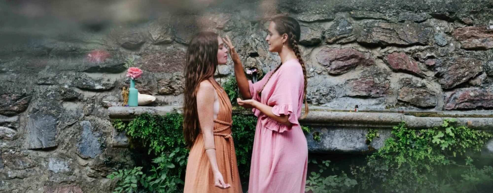 One woman anointing another’s brow at a mossy stone spring