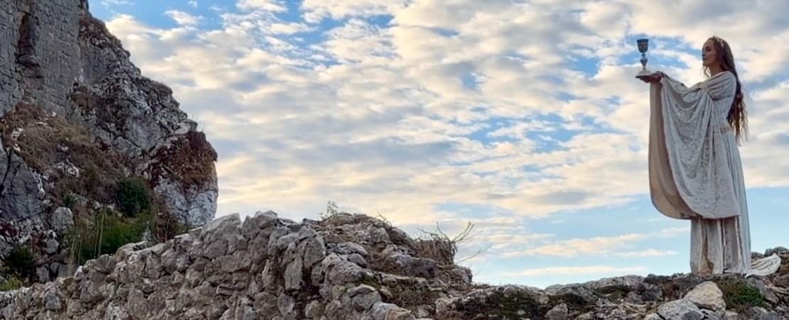A woman in white raising a chalice on a rocky ridge beneath a wide clouded sky