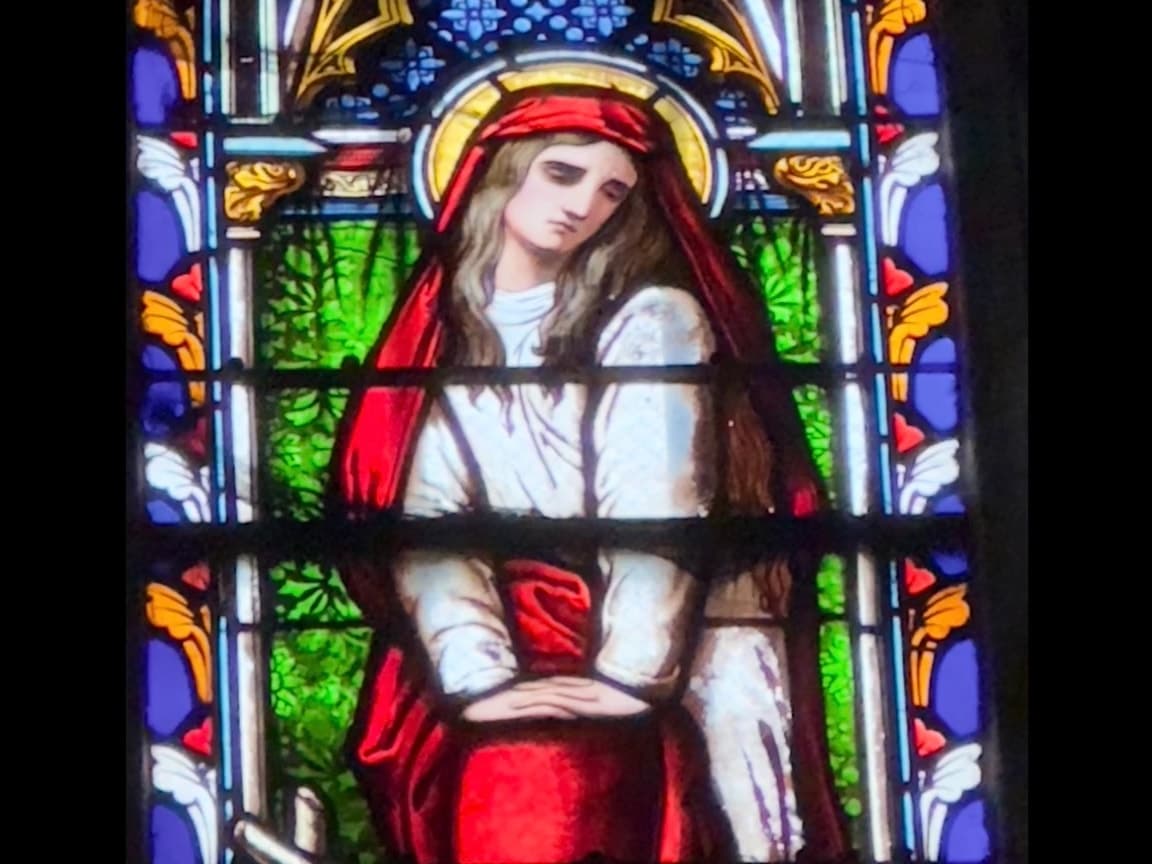 A stained-glass Magdalene veiled in red, her hands folded