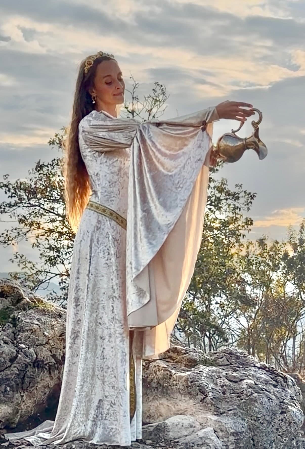 Dakota Chanel in white velvet on a rocky outcrop at sunset, pouring water from a brass ewer