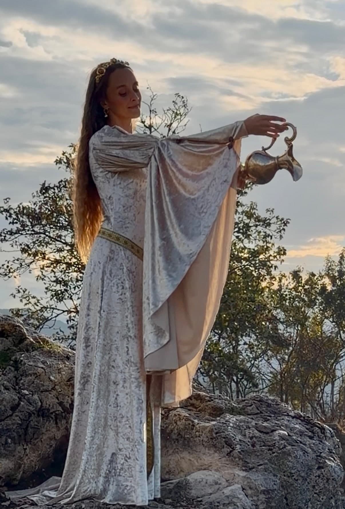 Dakota Chanel in white velvet on a rocky outcrop at sunset, pouring water from a brass ewer