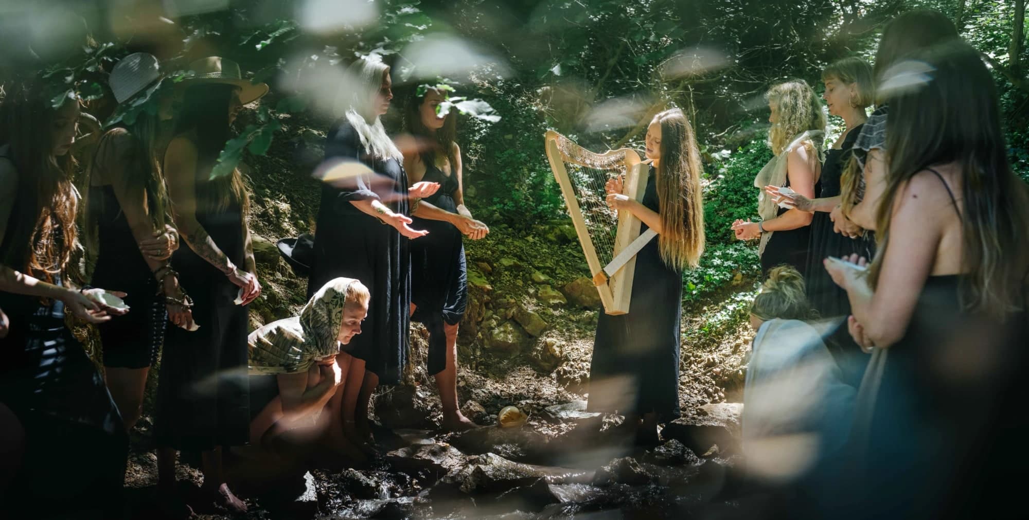 The sisterhood gathered at a forest stream, one woman playing the harp
